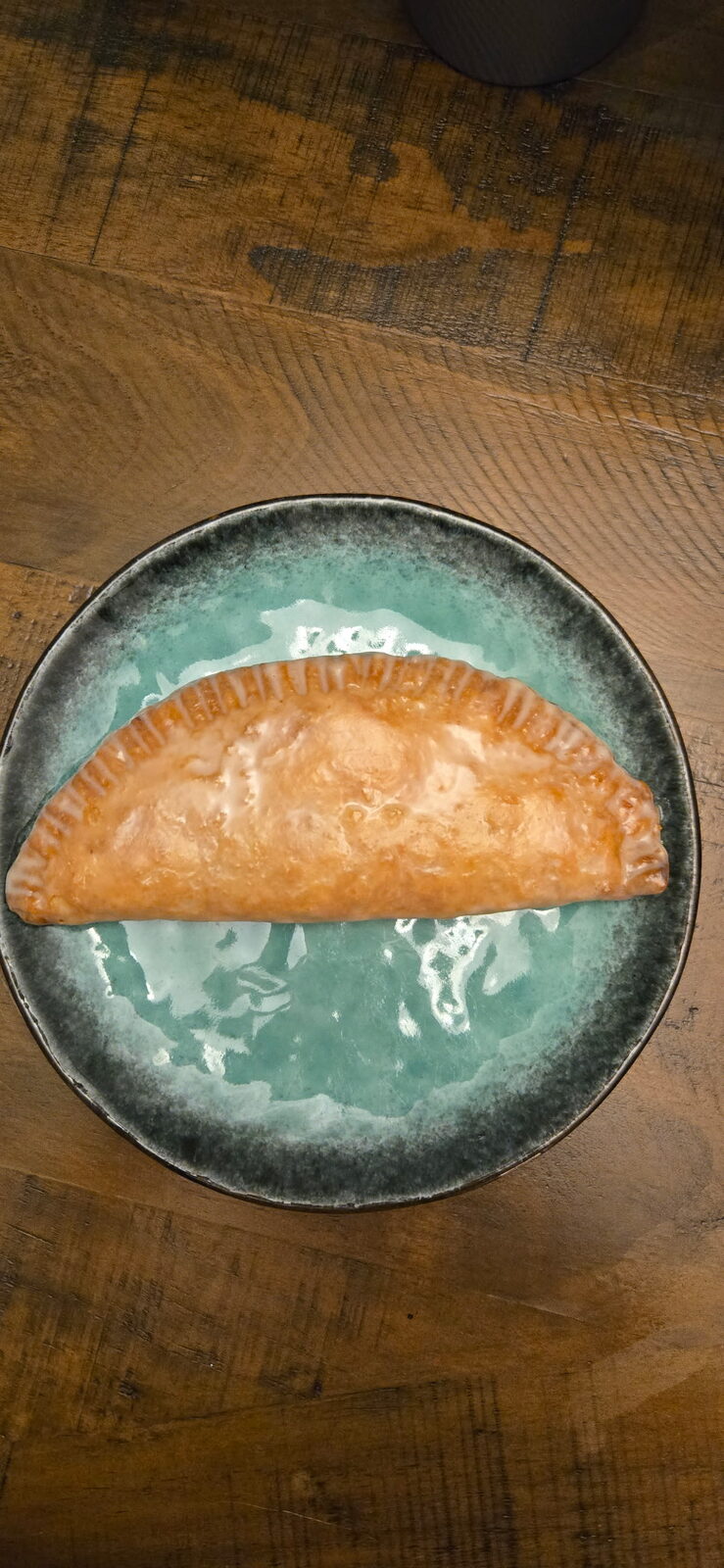 A golden hand-folded fried pie, crimped along the edge, resting on a teal ceramic plate on a dark wood table
