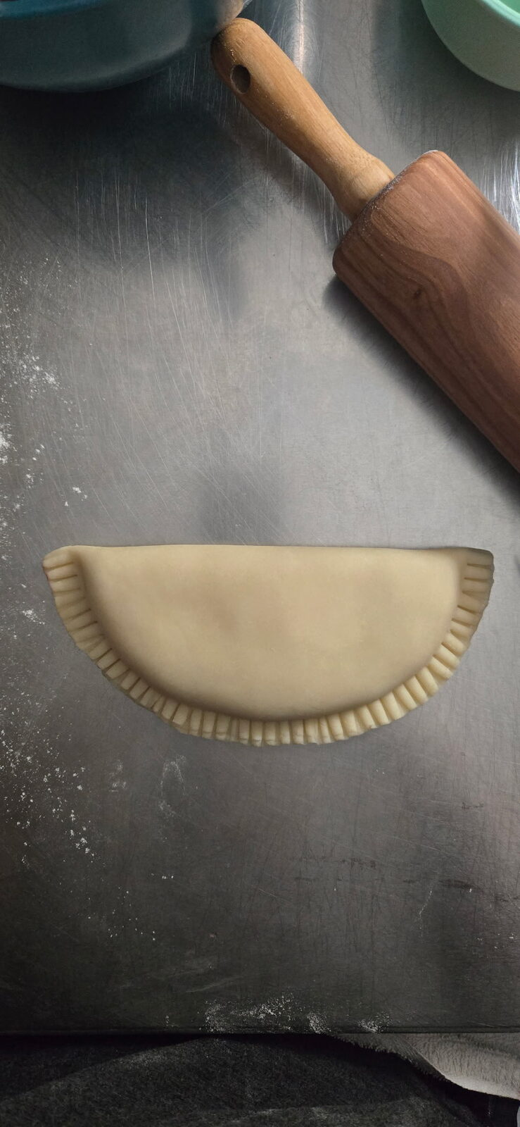 An uncooked pie crimped shut by hand on a floured steel counter, wooden rolling pin beside it